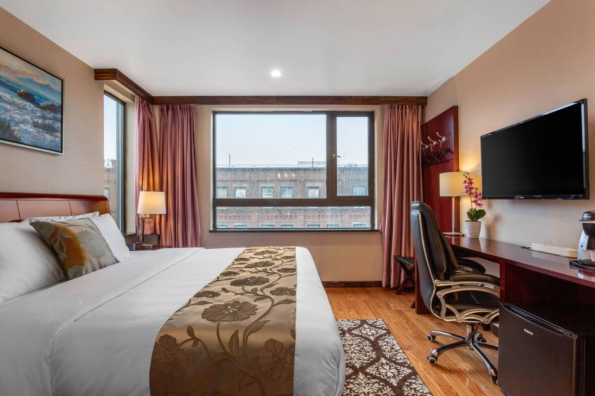 A hotel room with a large bed, a desk with a computer, a flat-screen TV, and a window with curtains.