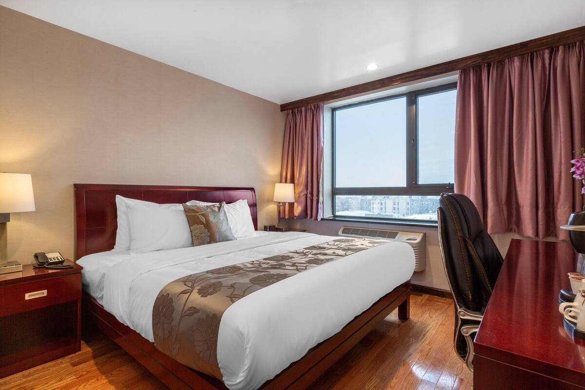 A hotel room with a large bed, two bedside tables with lamps, a desk with a chair, and a window with curtains.