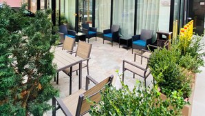 Exterior de un hotel con diseño moderno, que cuenta con un amplio muro de cristal, un área de estar al aire libre con sillas y mesas, y un jardín cuidado con plantas en macetas.