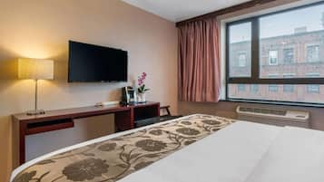Habitación de hotel con cama, televisor de pantalla plana, escritorio con lámpara, ventana con cortinas y vista a edificios.