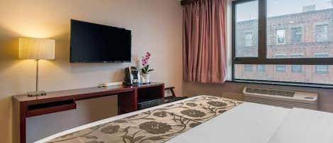 A hotel room with a bed, a flat-screen TV, a desk with a lamp, a window with curtains, and a view of buildings outside.