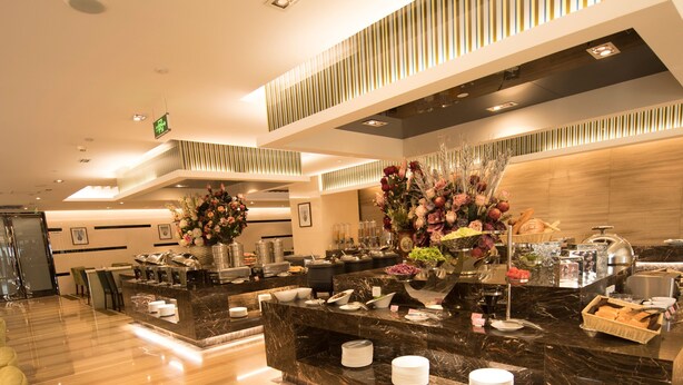 Een moderne hotel lobby met marmeren vloeren, een receptiebalie met een bloemstuk en een buffetruimte.