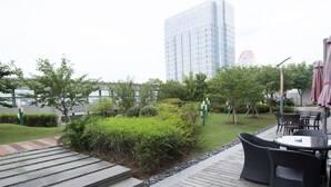 Een houten terras met buitenmeubilair, een gazon met struiken en een hoog gebouw op de achtergrond.