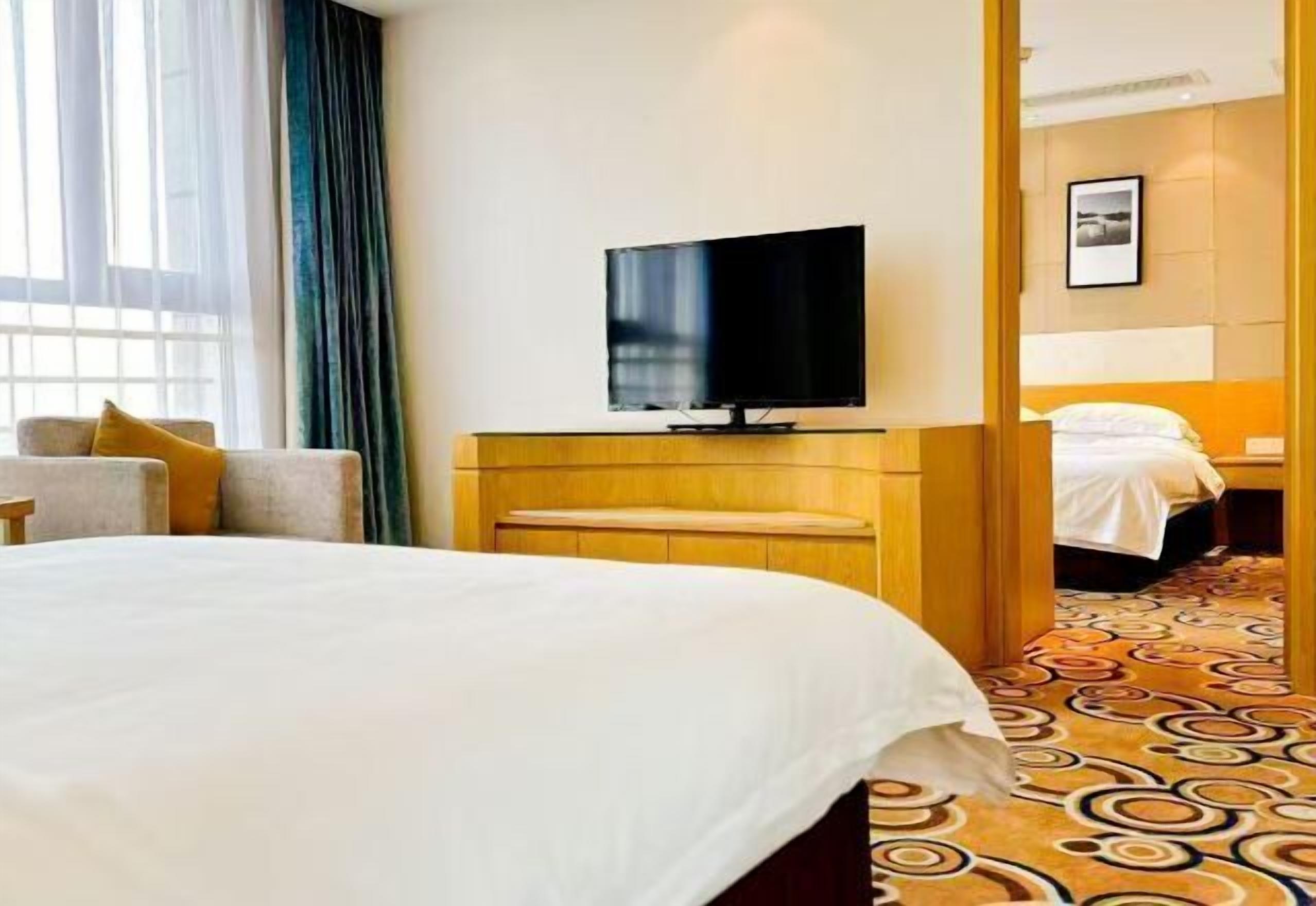 A hotel room with a large bed, a television on a wooden stand, a desk, and a patterned carpet.