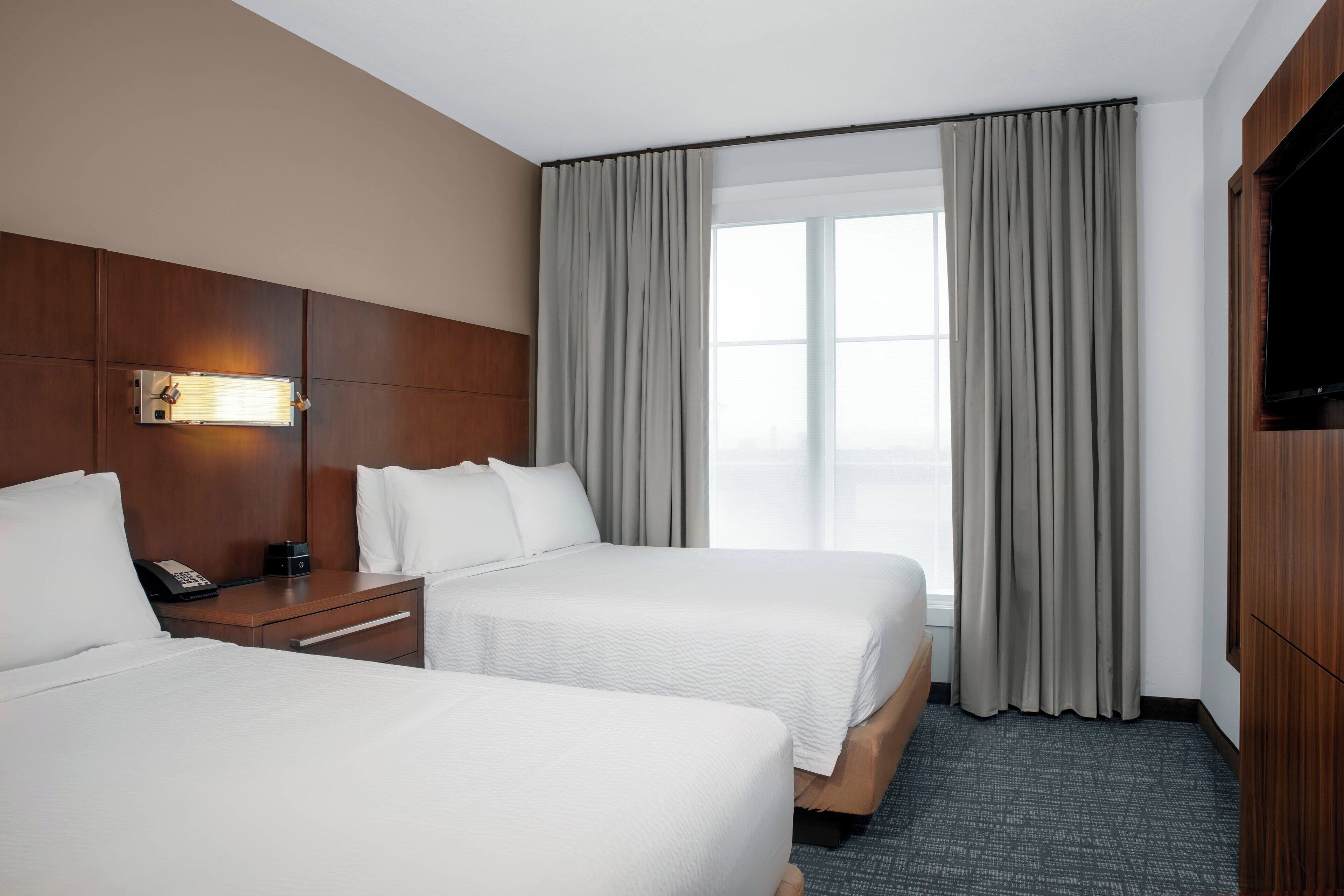 Une chambre d’hôtel avec deux lits, une tête de lit en bois, une table de chevet avec un téléphone et une grande fenêtre avec des rideaux.
