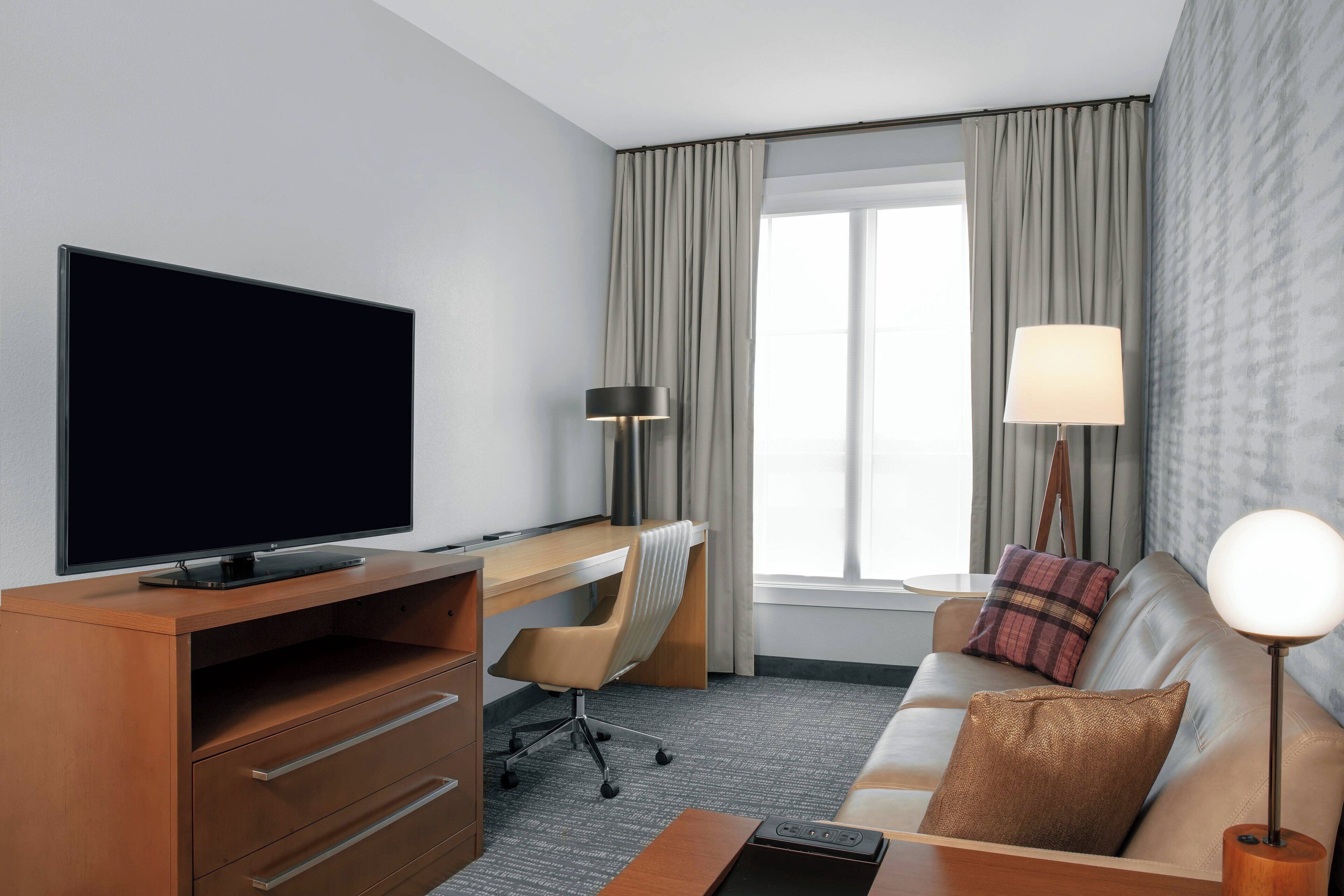 A hotel room with a flat-screen TV, a desk with a chair, a sofa with a lamp, and a window with curtains.