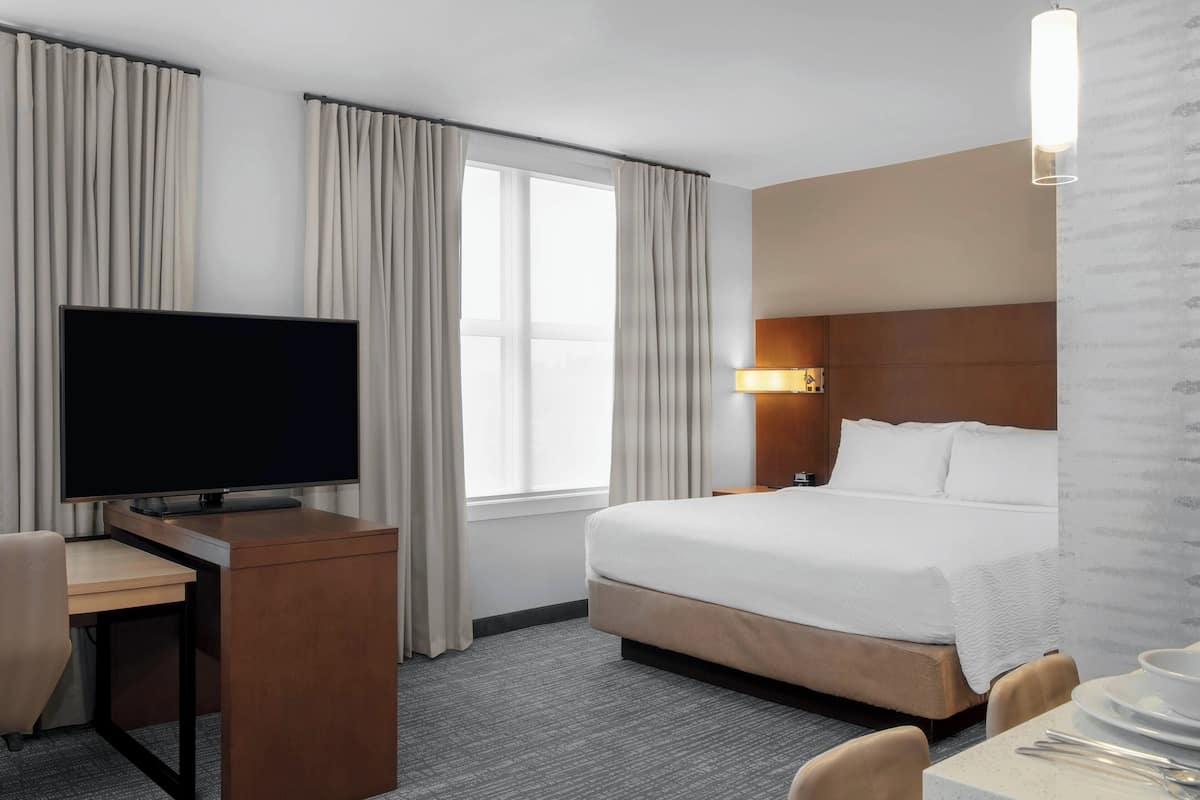 A hotel room with a bed, a desk with a television, a lamp, and a window with curtains.