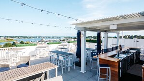 Une terrasse extérieure avec des tables et des chaises, un espace bar et une vue sur l’eau et le paysage urbain.