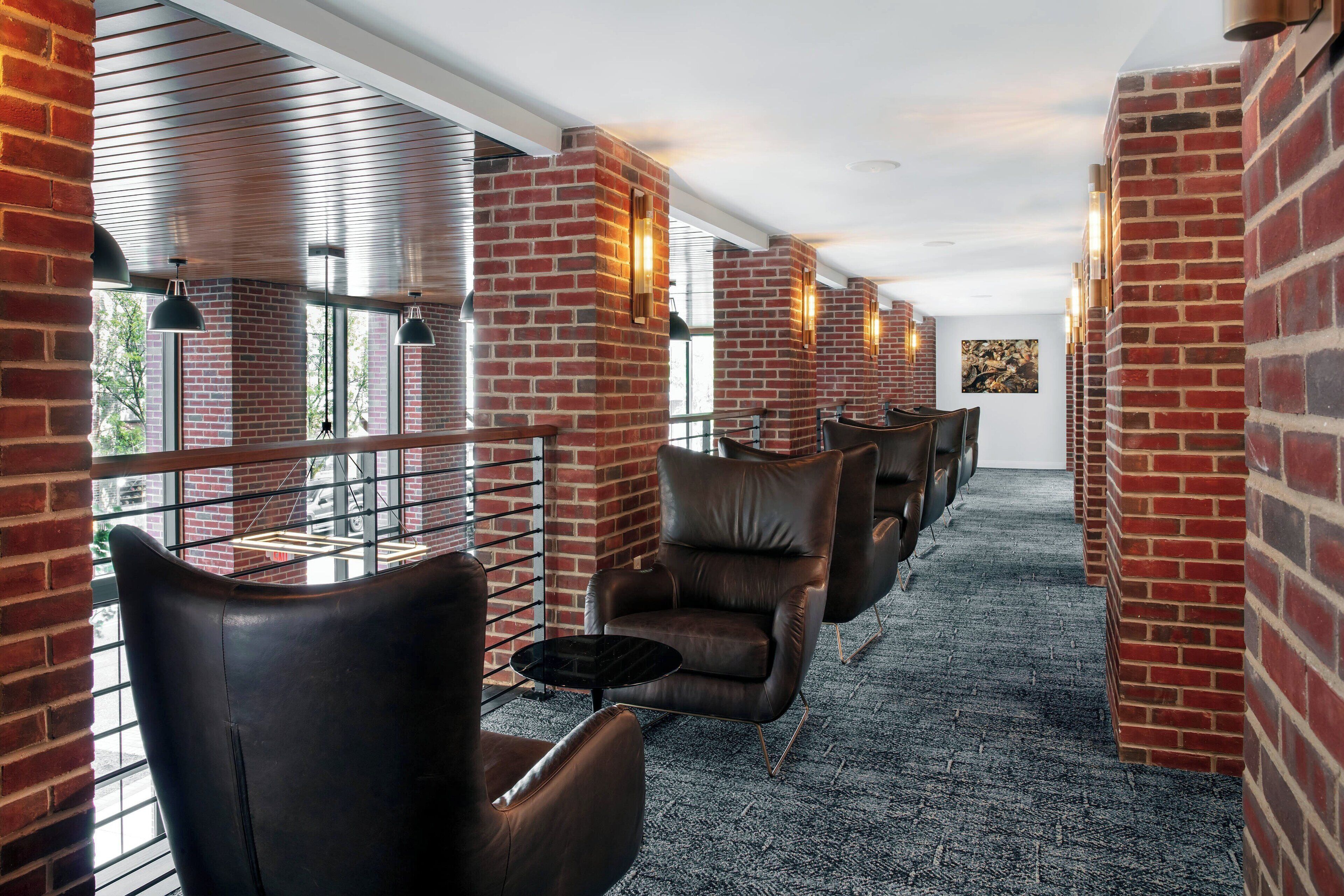 A corridor with brick walls, leather chairs, and tables, leading to a door.