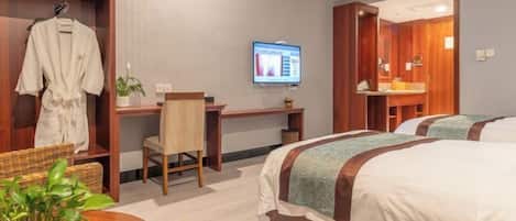 Quarto de hotel com duas camas, uma escrivaninha, uma cadeira, uma televisão e casa de banho com espelho.