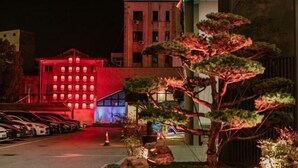 A building with a red-lit facade, a parking lot with cars, and a well-maintained garden with trees. - Songling Hotel (Suzhou)