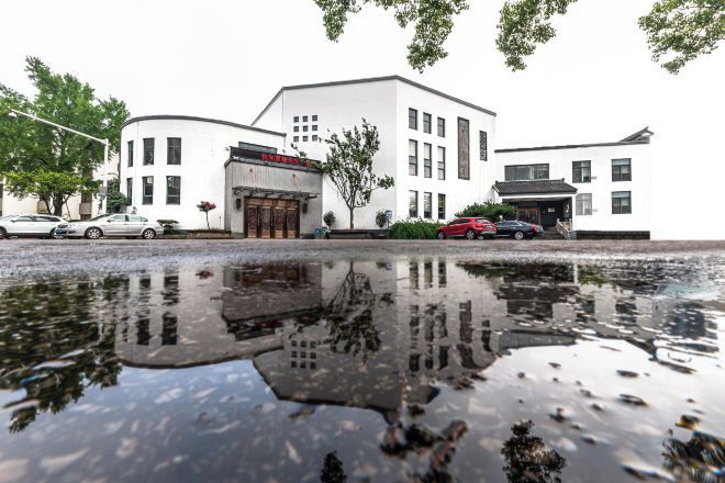 A white building with a reflection in a puddle, a red car, and a tree.