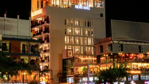 Un hôtel à plusieurs étages, reconnaissable à son grand panneau « MENIO HOTEL », doté de nombreux balcons et abritant un café au rez-de-chaussée.