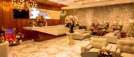 A hotel reception area with a modern design, featuring a wooden reception desk, a stone wall, and multiple seating areas with floral arrangements.