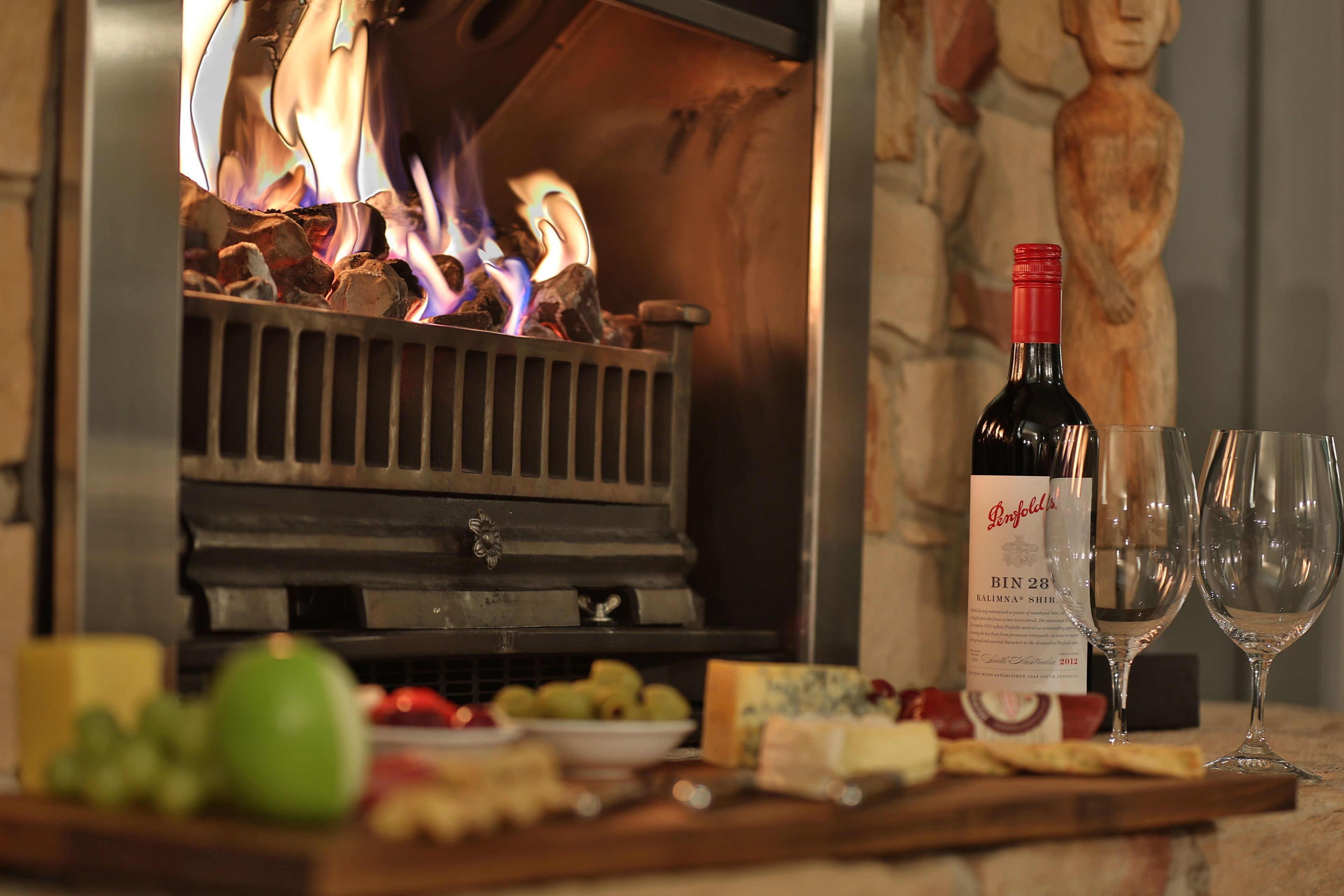 A fireplace with burning logs, a bottle of wine, two glasses, and a cheese board with fruits and crackers.