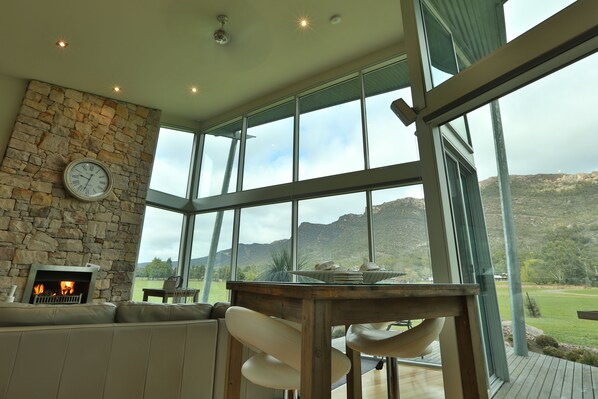 A spacious room with a stone fireplace, large windows offering a view of the outdoors, and a wooden table with chairs. - Ironstone Romantic Couples Retreat (Halls Gap)