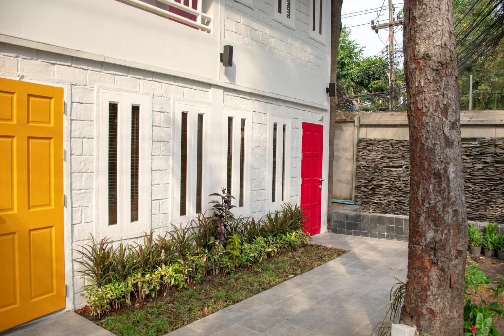 A building with white brick walls, a yellow door, and a red door. There is a small garden with plants in front of the building.