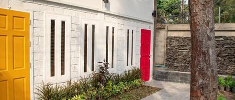 A building with white brick walls, a yellow door, and a red door. There is a small garden with plants in front of the building.