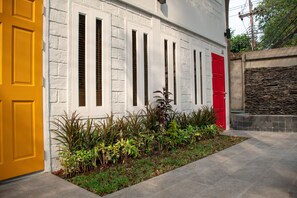 A building with a yellow door, white brick walls, and a red door. There is a small garden with various plants in front of the building. - Lazy Sandals Koh Samed (Rayong)