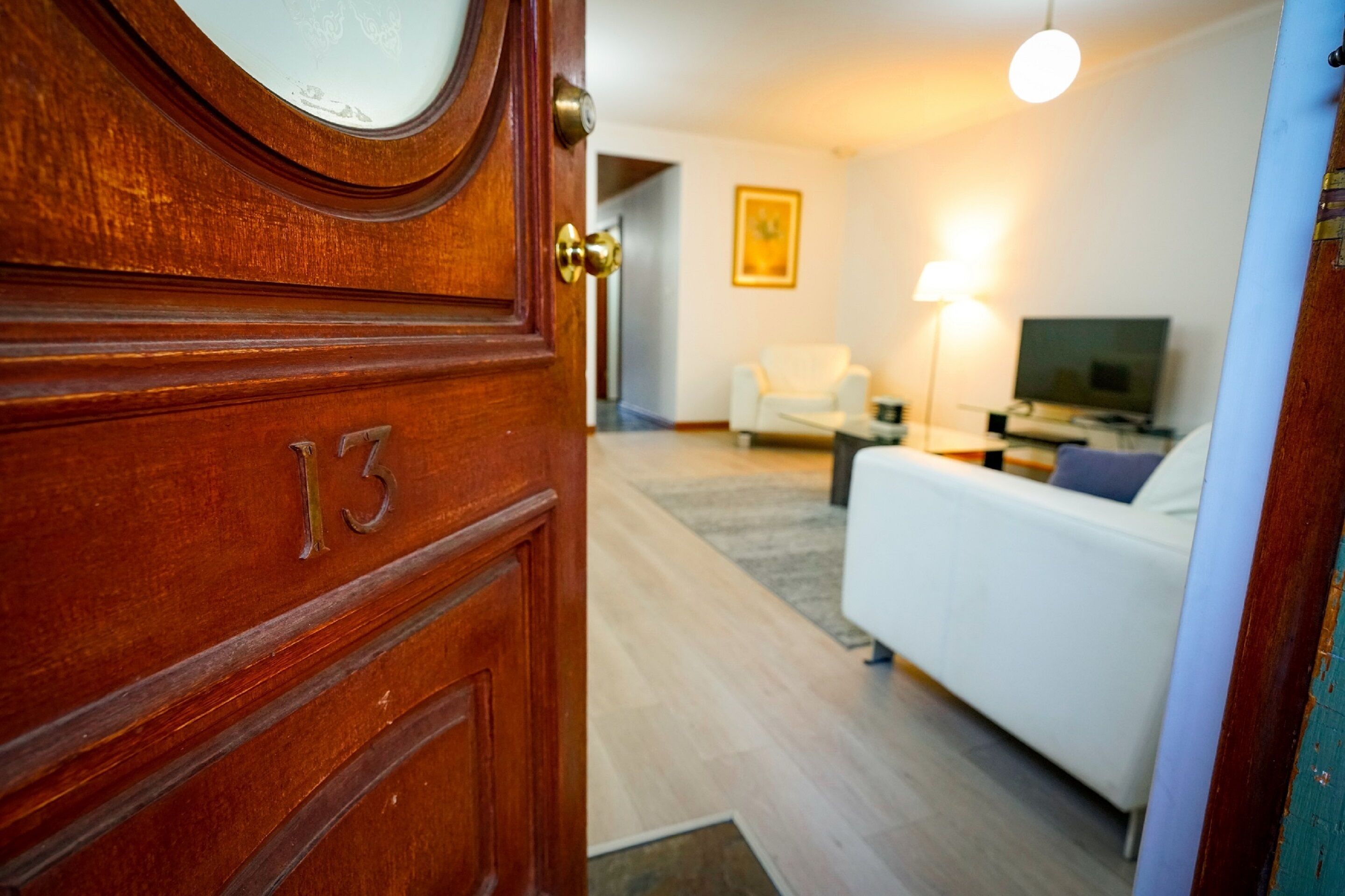 A wooden door with the number 13, leading into a room with a bed, a white chair, a lamp, and a television.