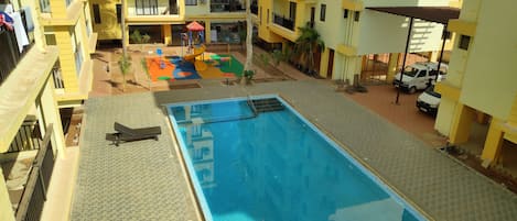 A swimming pool with a tiled deck, surrounded by yellow buildings with balconies and a children's play area.