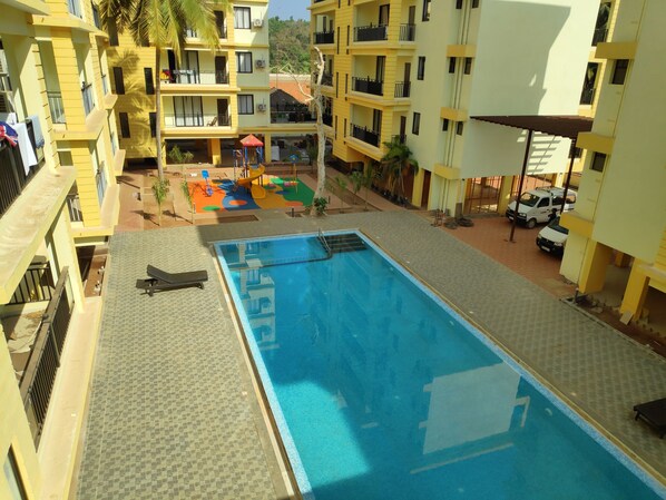 A swimming pool with a tiled deck, surrounded by yellow buildings with balconies and a children's play area.