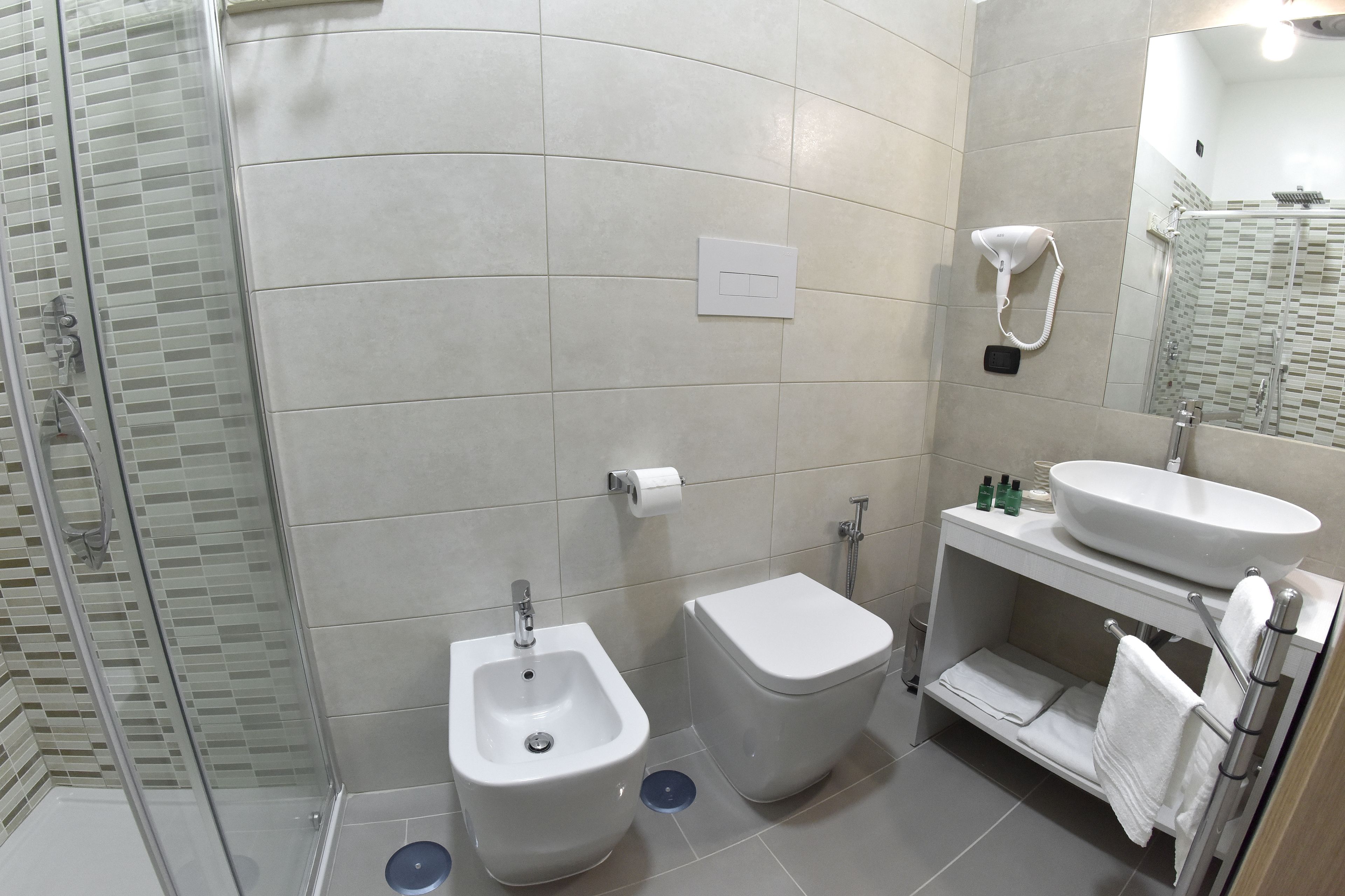 Ein modernes Badezimmer mit einer einachigen Duschkabine, einem Bidet, einer Toilette, einem Waschbecken mit Spiegel und einem Handtuchhalter.