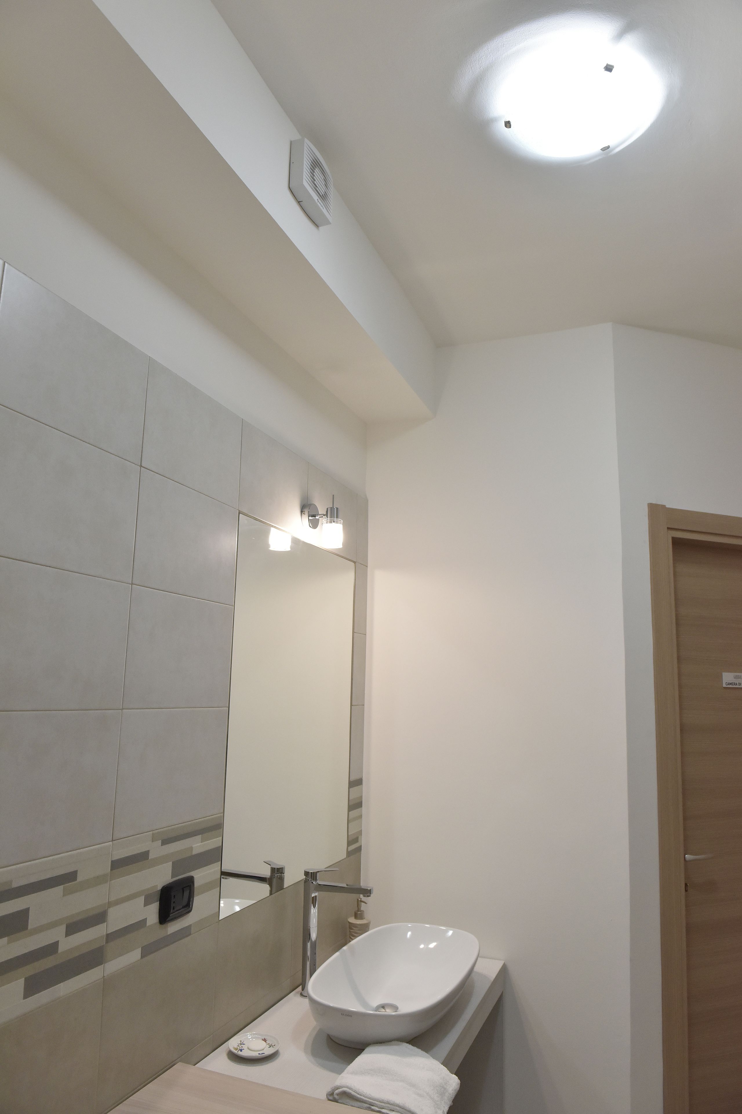 Une salle de bain moderne avec un grand miroir, un lavabo sur colonne et un luminaire au plafond.