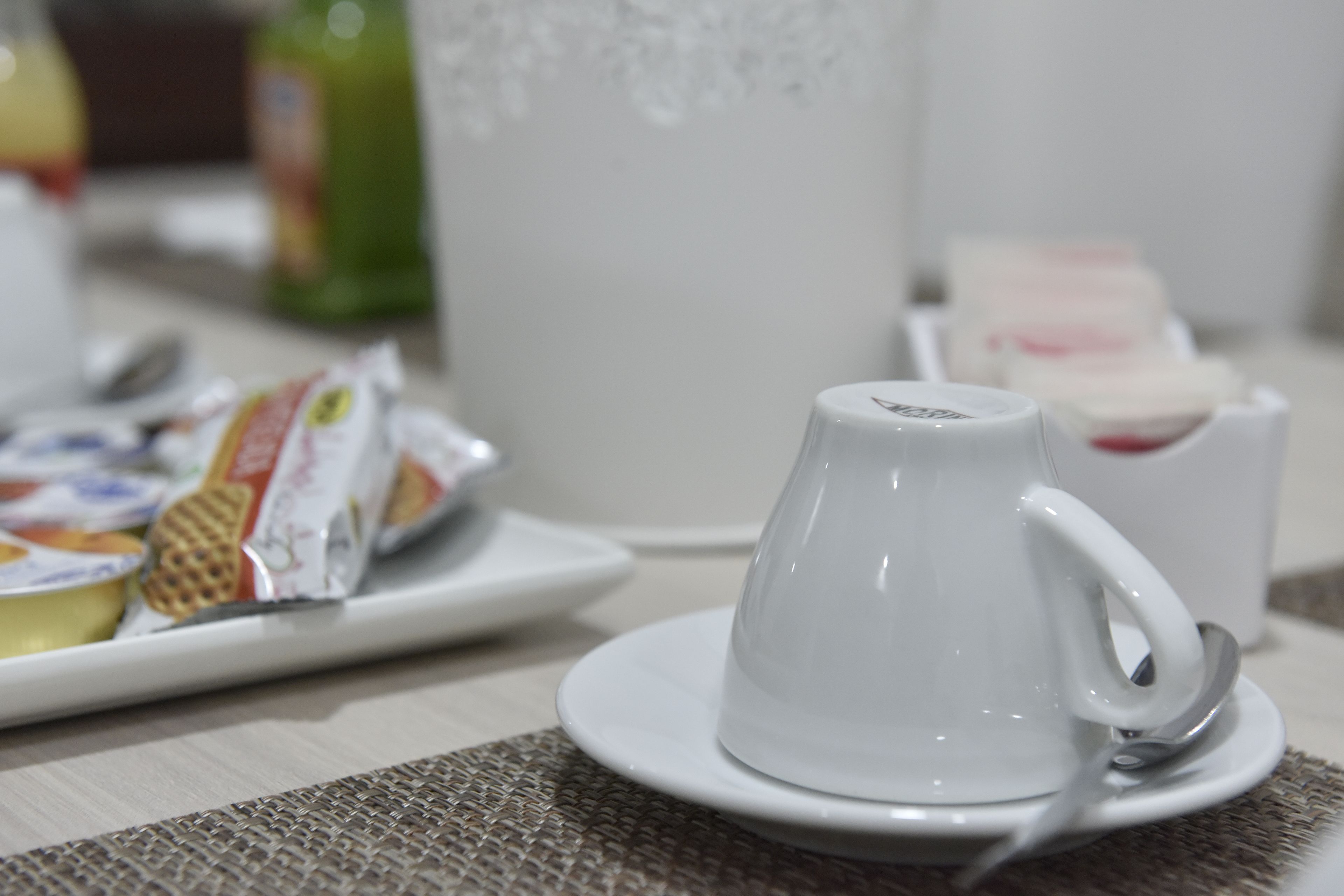 Una taza de cerámica blanca sobre un plato con una cuchara, un plato con porciones de comida, y una botella verde al fondo.