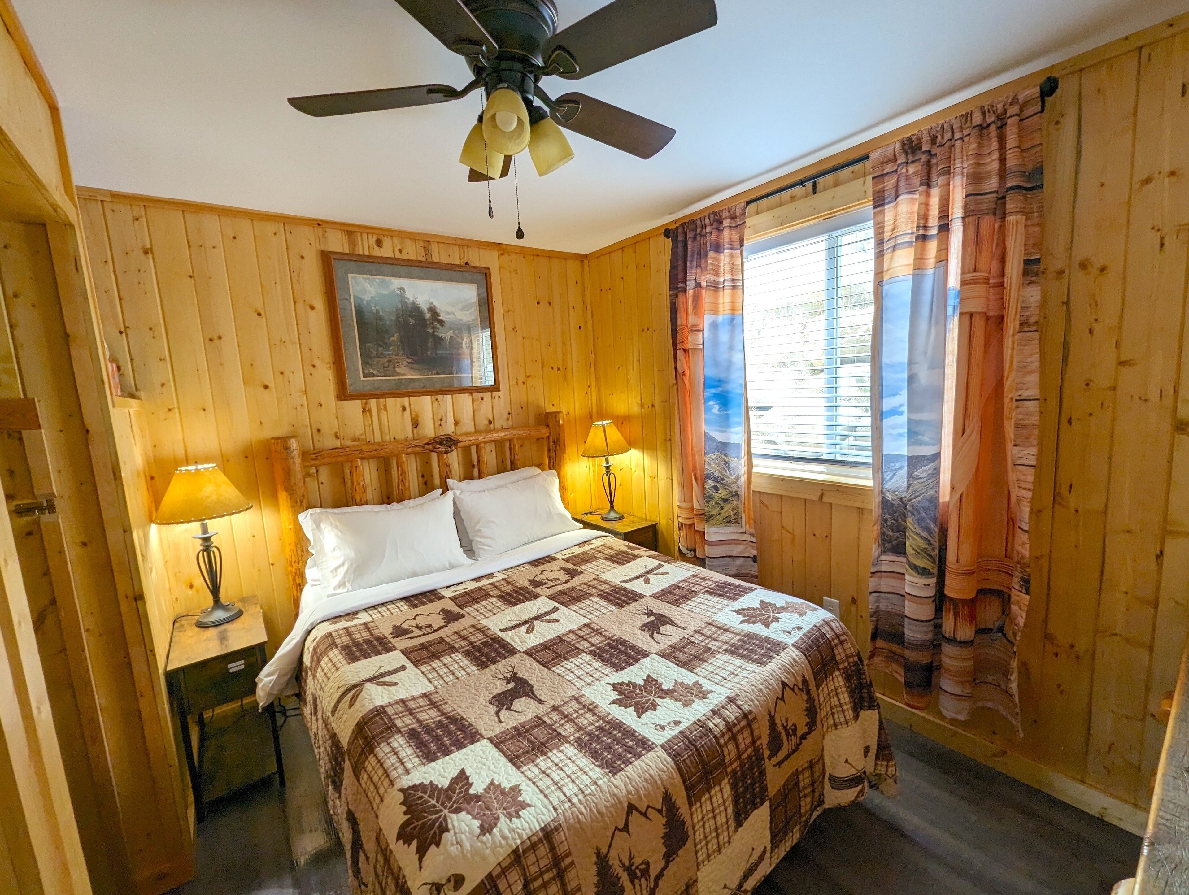 A wooden-paneled room with a bed, two bedside lamps, a ceiling fan, and a window with curtains.