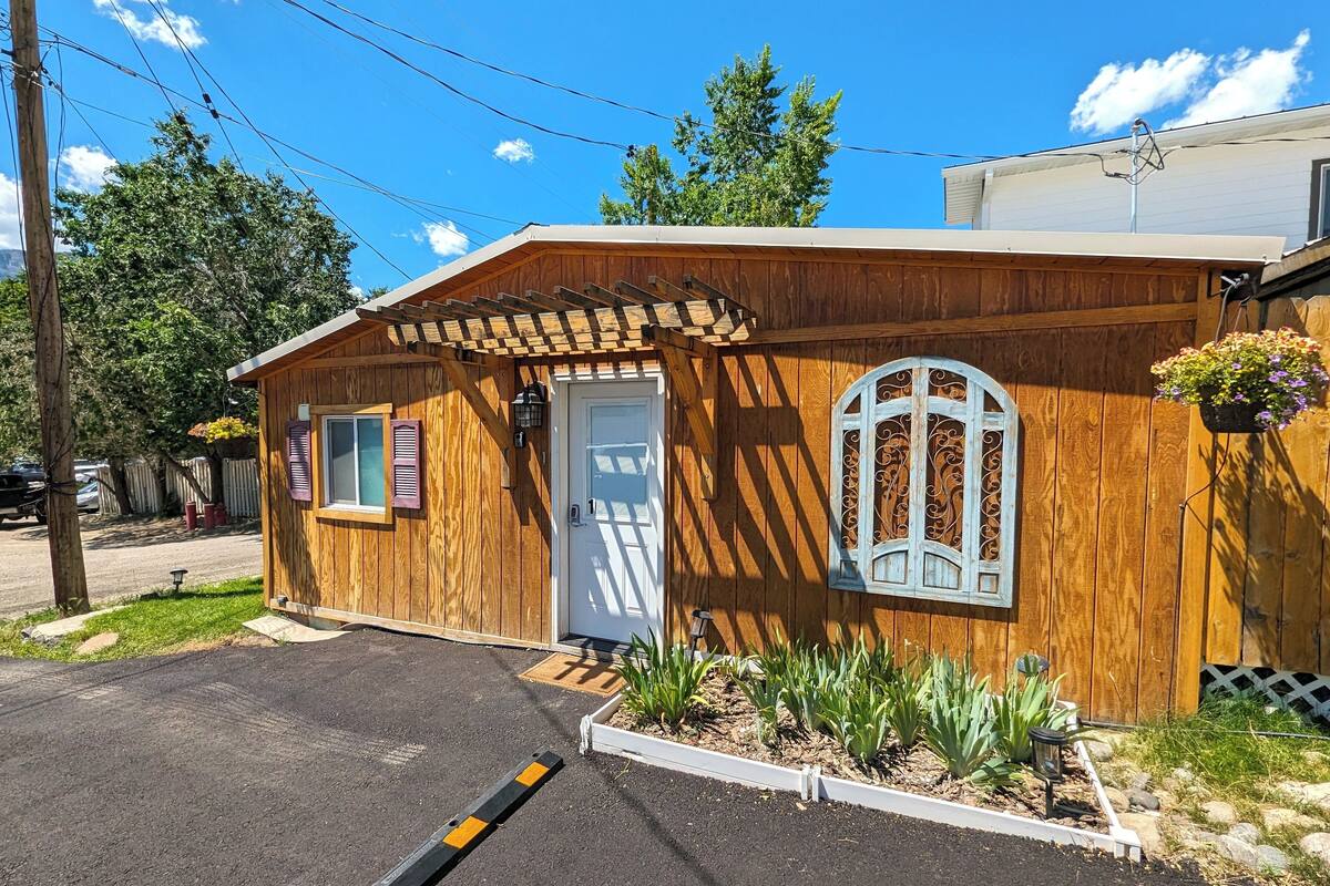 A wooden cabin with a white door, a small porch, and a garden area with plants and a hanging flower pot.