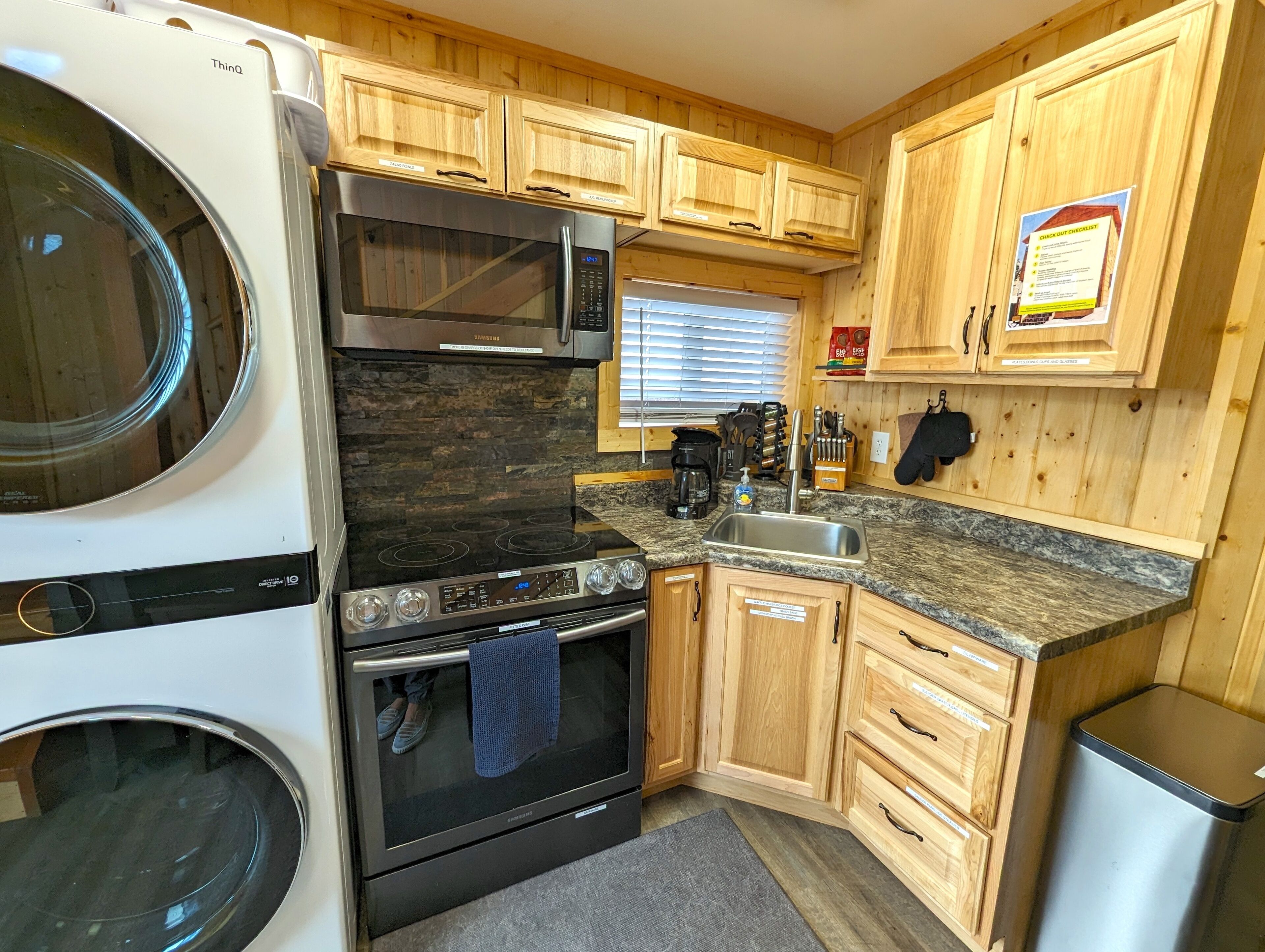 A compact kitchen with a built-in washer and dryer, stainless steel appliances, wooden cabinets, granite countertops, and a check-out checklist on the wall.