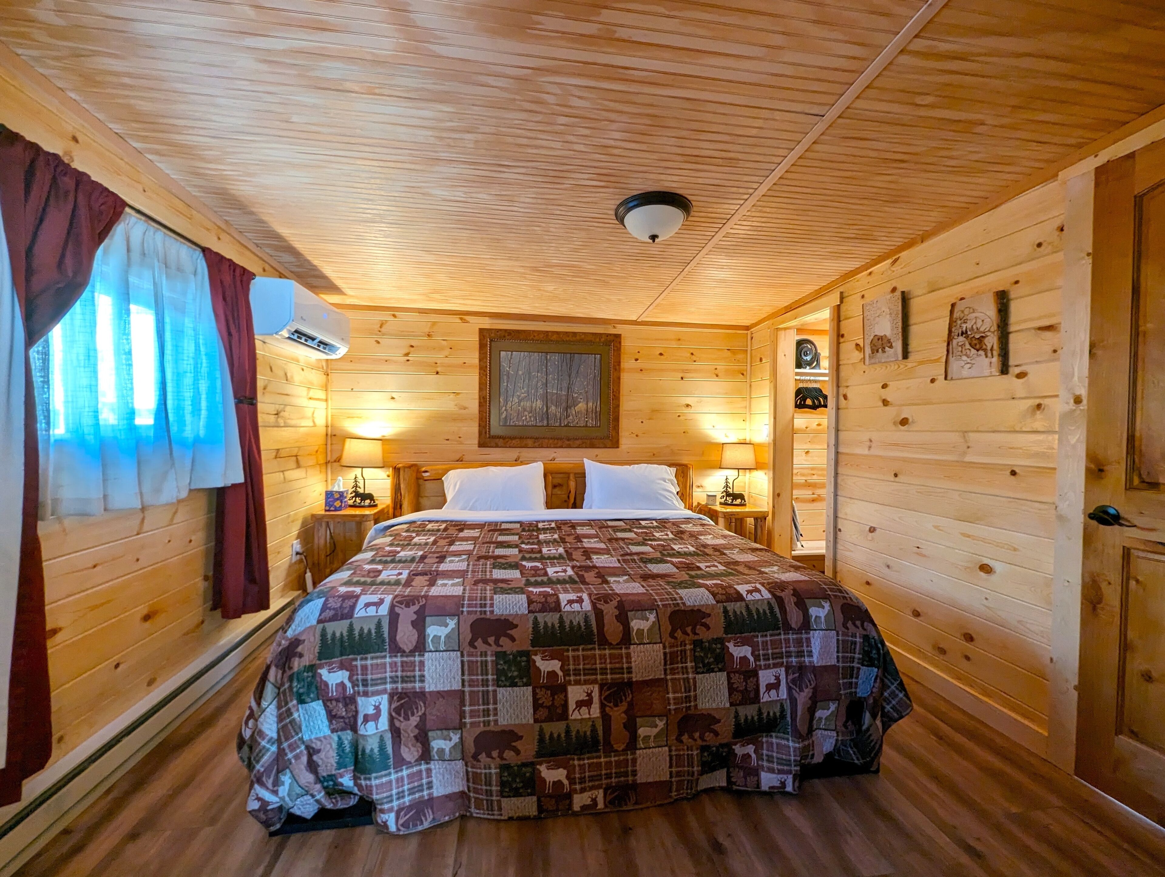 A wooden cabin room with a bed, two bedside tables, a wall-mounted air conditioner, and a window with curtains.