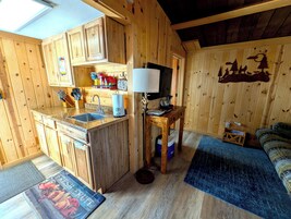 Una cocina compacta con armarios de madera, encimera de granito y un lavamanos. Hay una pequeña mesa con una lámpara y una alfombra azul en el suelo.