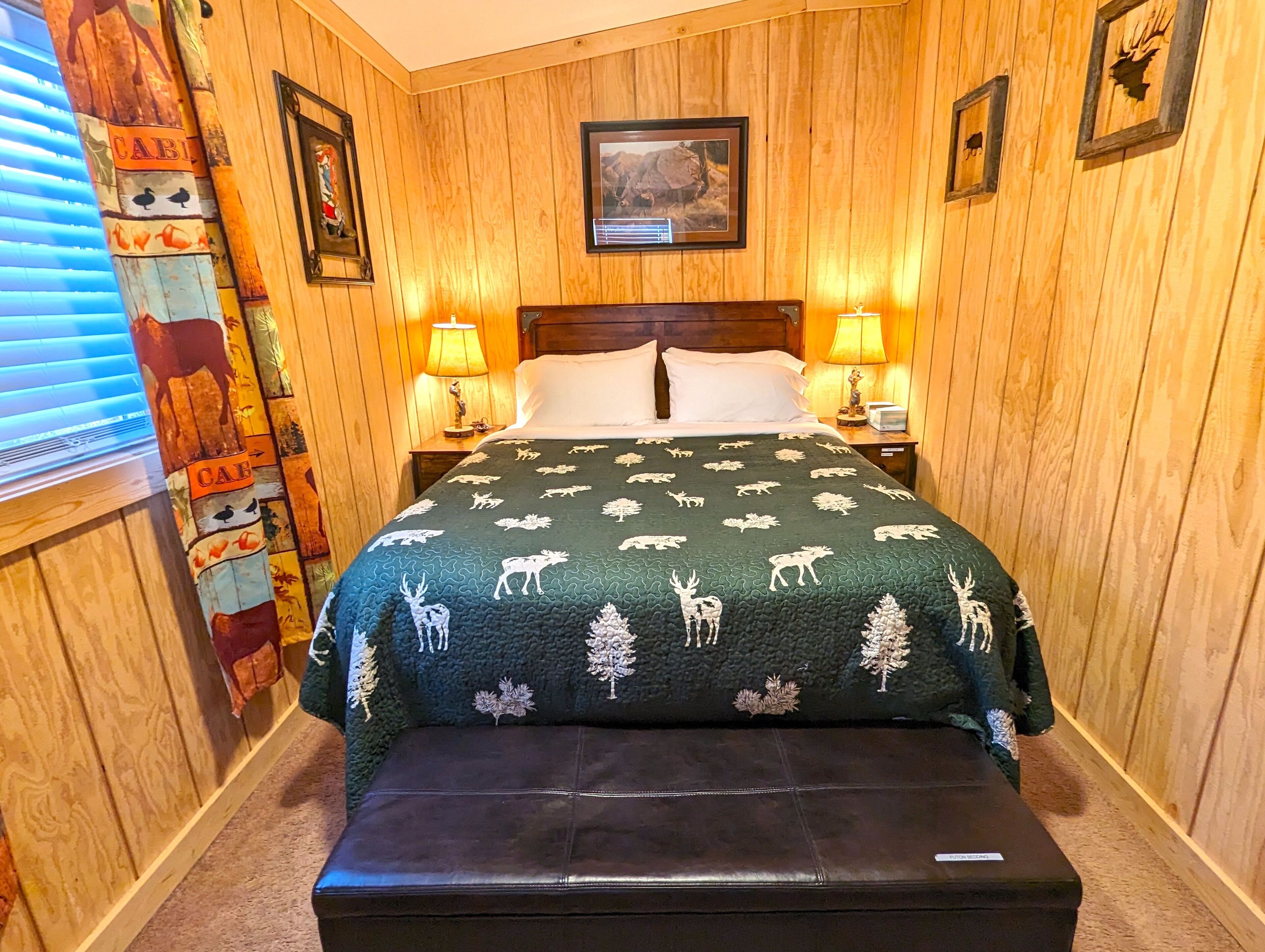A room with wooden walls, a bed with a green blanket featuring animal prints, two bedside lamps, and a wooden bench.