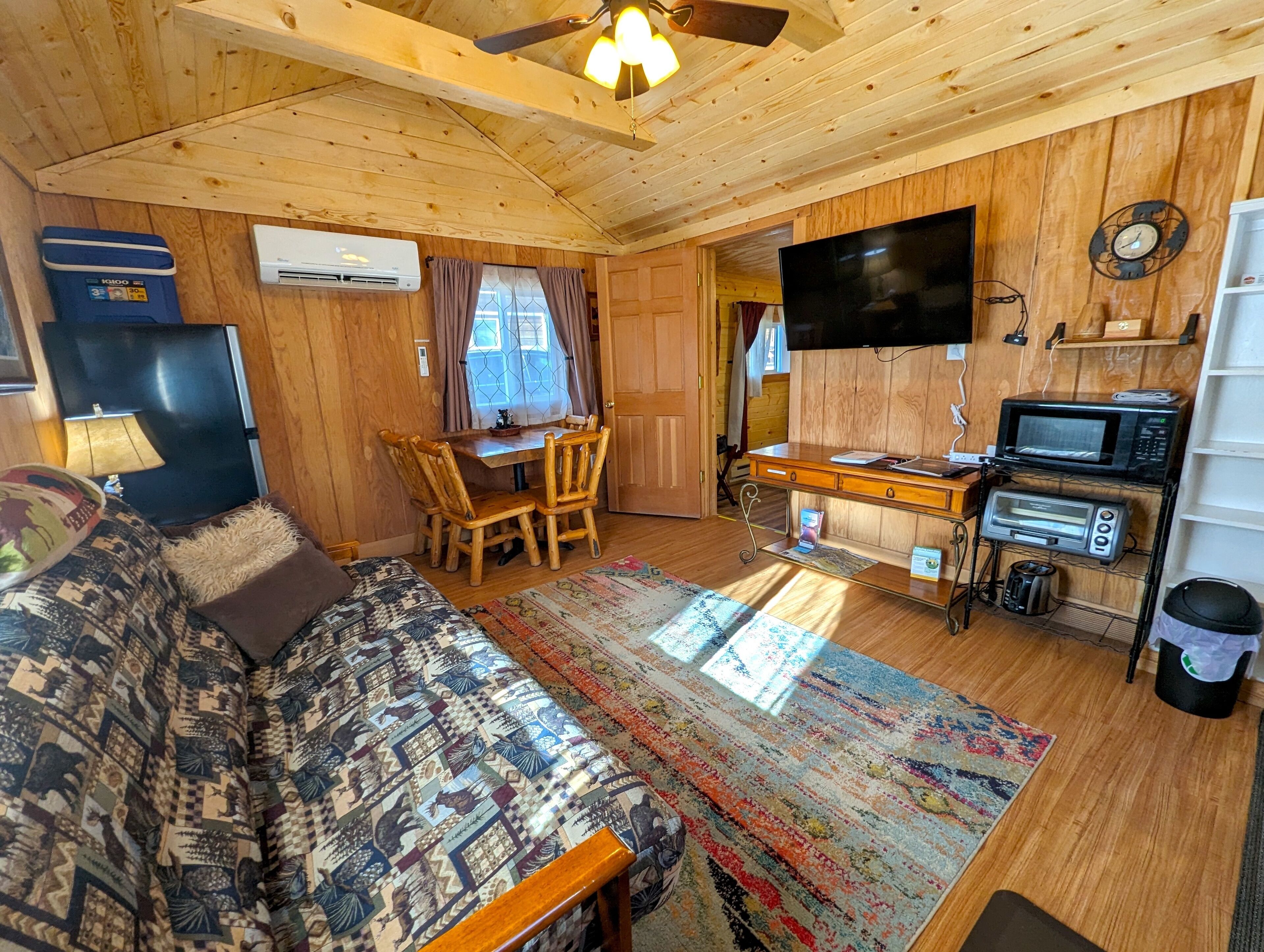A cozy cabin interior with a wooden ceiling, a sofa, a dining table with chairs, a TV, a microwave, a toaster, and a small kitchen area.