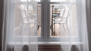 A room with a large window covered by sheer curtains, revealing outdoor furniture and a view of a building across the street.