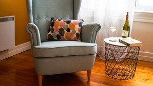A light blue armchair with a multicolored pillow, a round metal side table with a wine bottle and a book, and a window with sheer curtains.