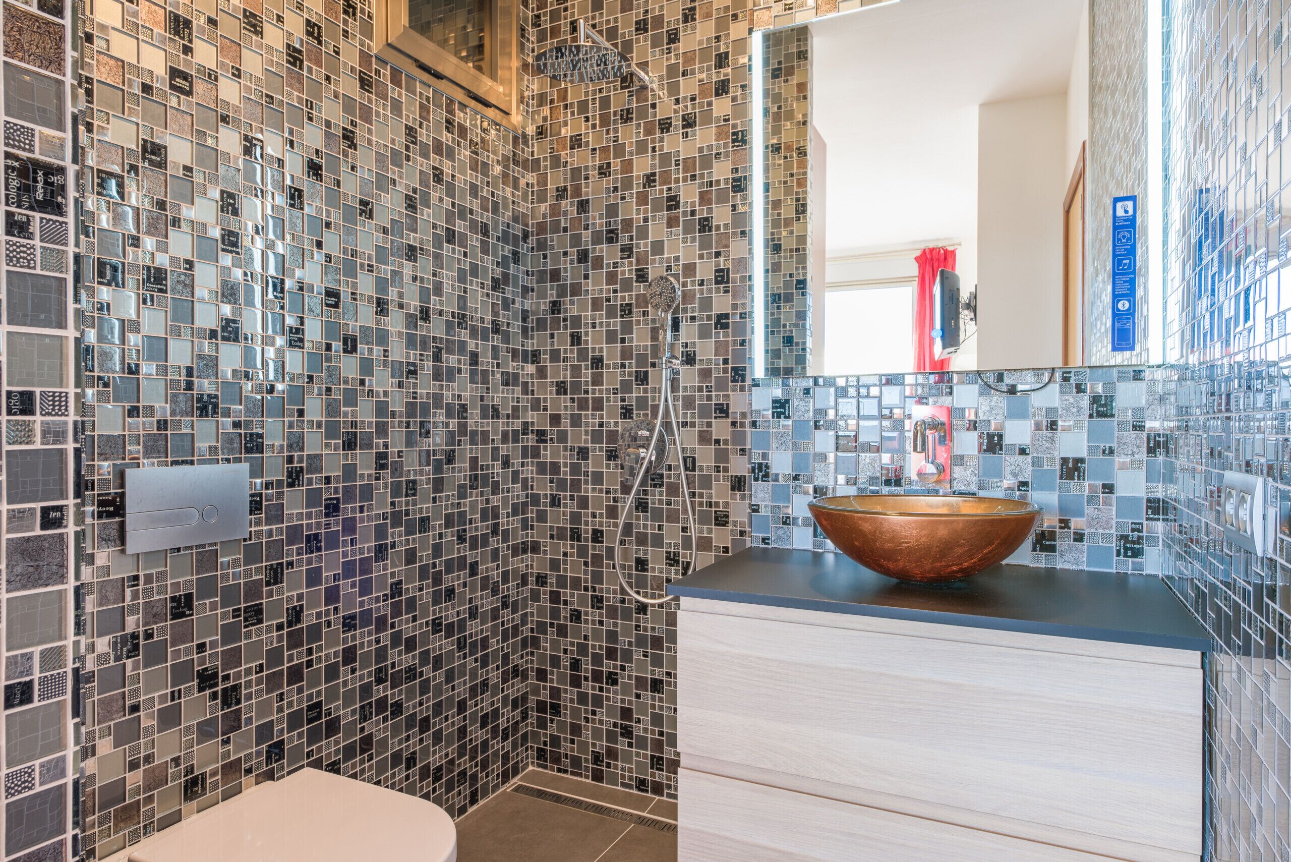 Une salle de bain moderne avec des murs recouverts de mosaïques, un lavabo en forme de bol en bois et une douche dotée d’un pommeau de douche à main.