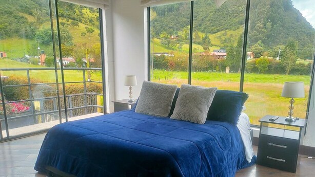 A bedroom with a large bed, a nightstand, a lamp, and a view of a green landscape through large windows. - Comfort Campestre (Tabio)