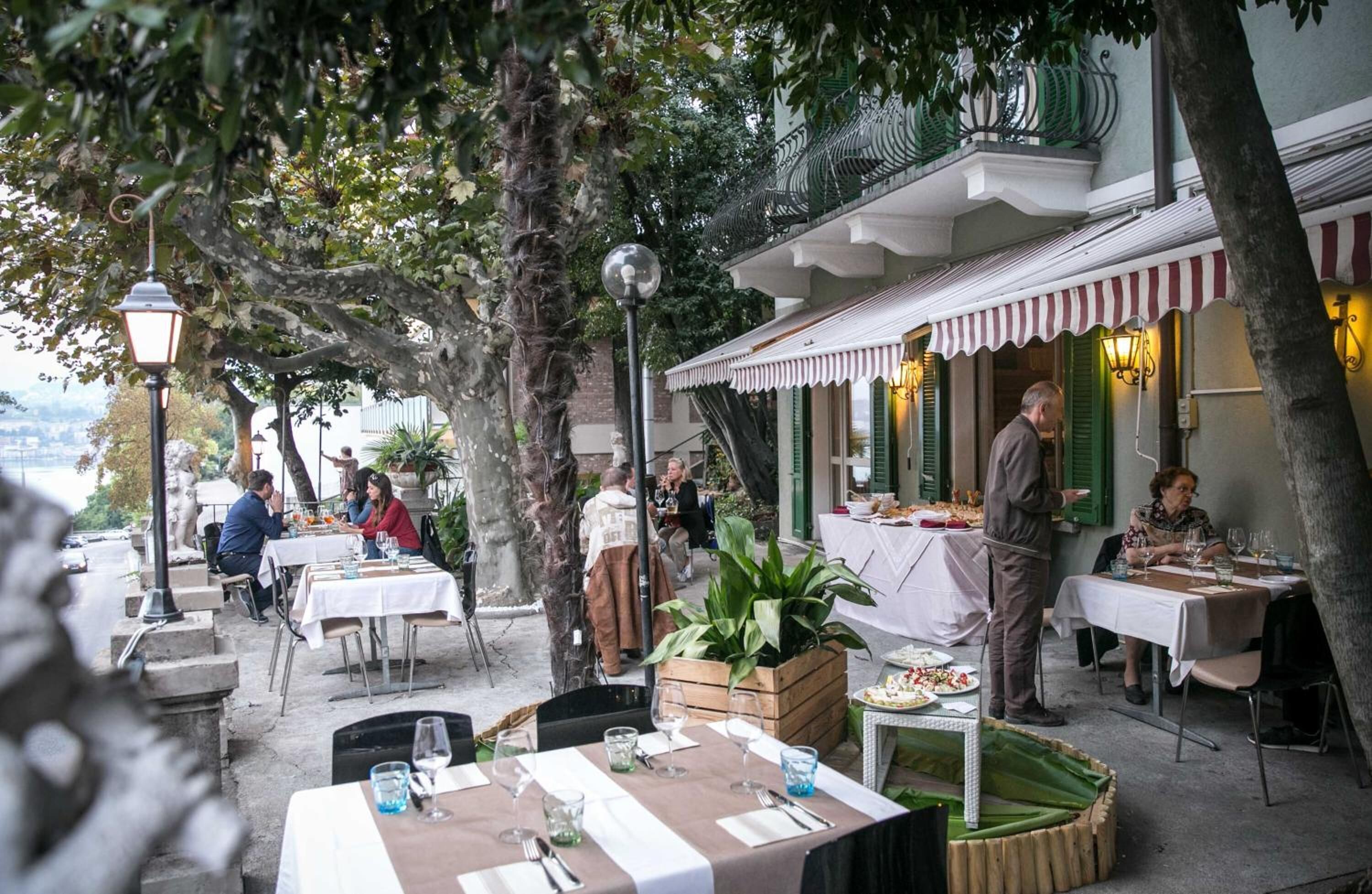 An outdoor dining area with tables and chairs, surrounded by trees and a building with a striped awning.