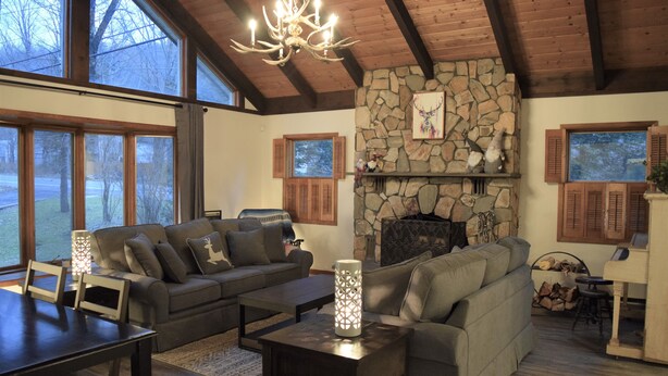 A spacious living room with a stone fireplace, wooden ceiling beams, and large windows offering a view of the outdoors.