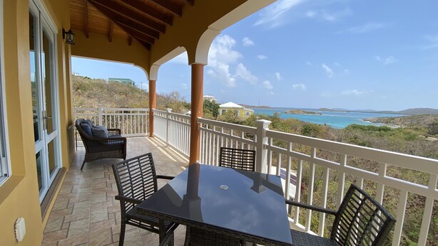 Ein Balkon mit Tisch und Stühlen, der auf eine Küstenlandschaft mit Blick auf das Meer und sanfte Hügel blickt.
