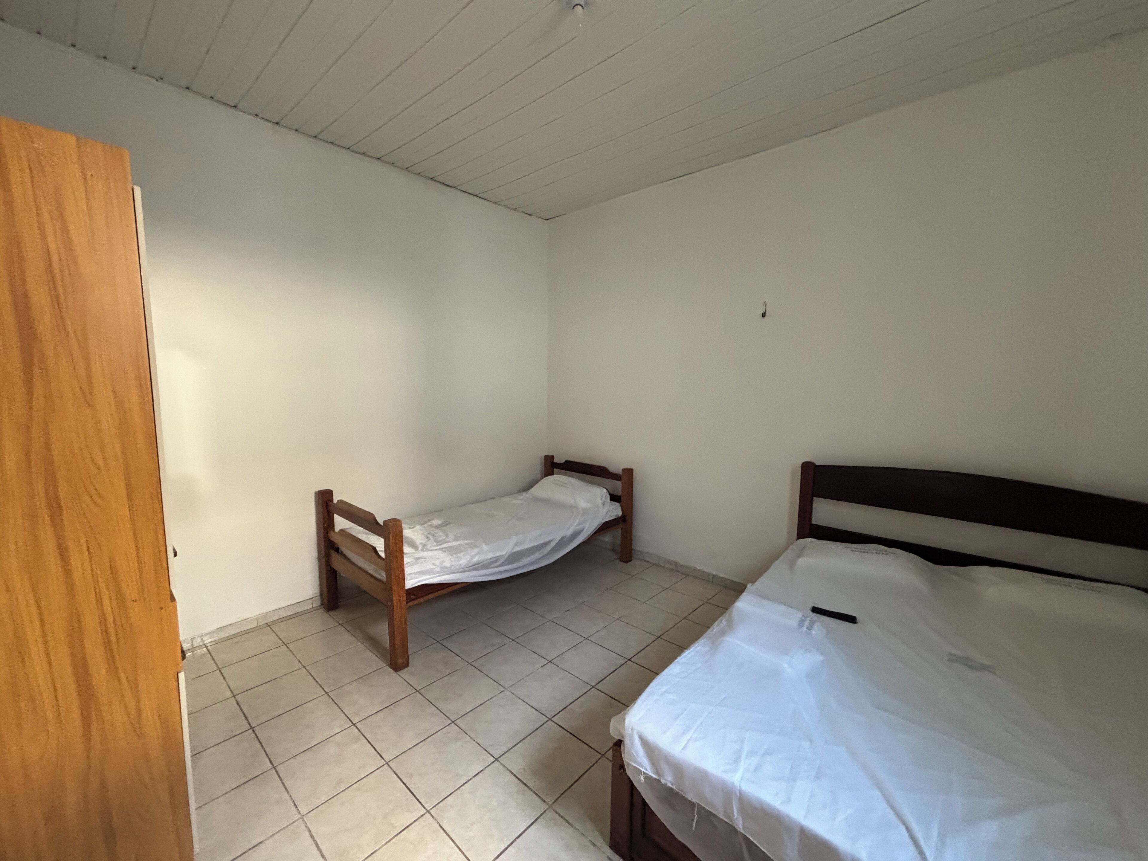 Um quarto pequeno e simples, com duas camas de solteiro, uma porta de madeira e chão de azulejos.