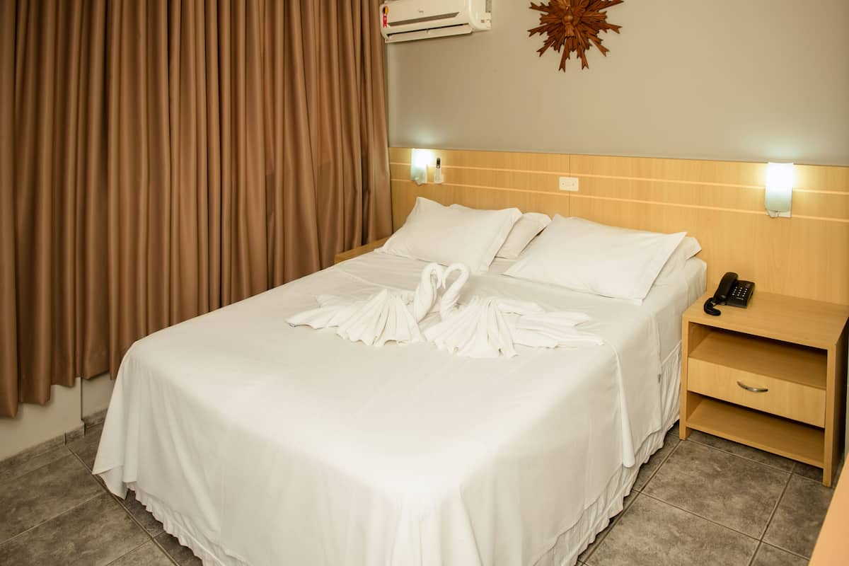 A hotel room with a neatly made bed, a bedside telephone, and a wall-mounted air conditioner.