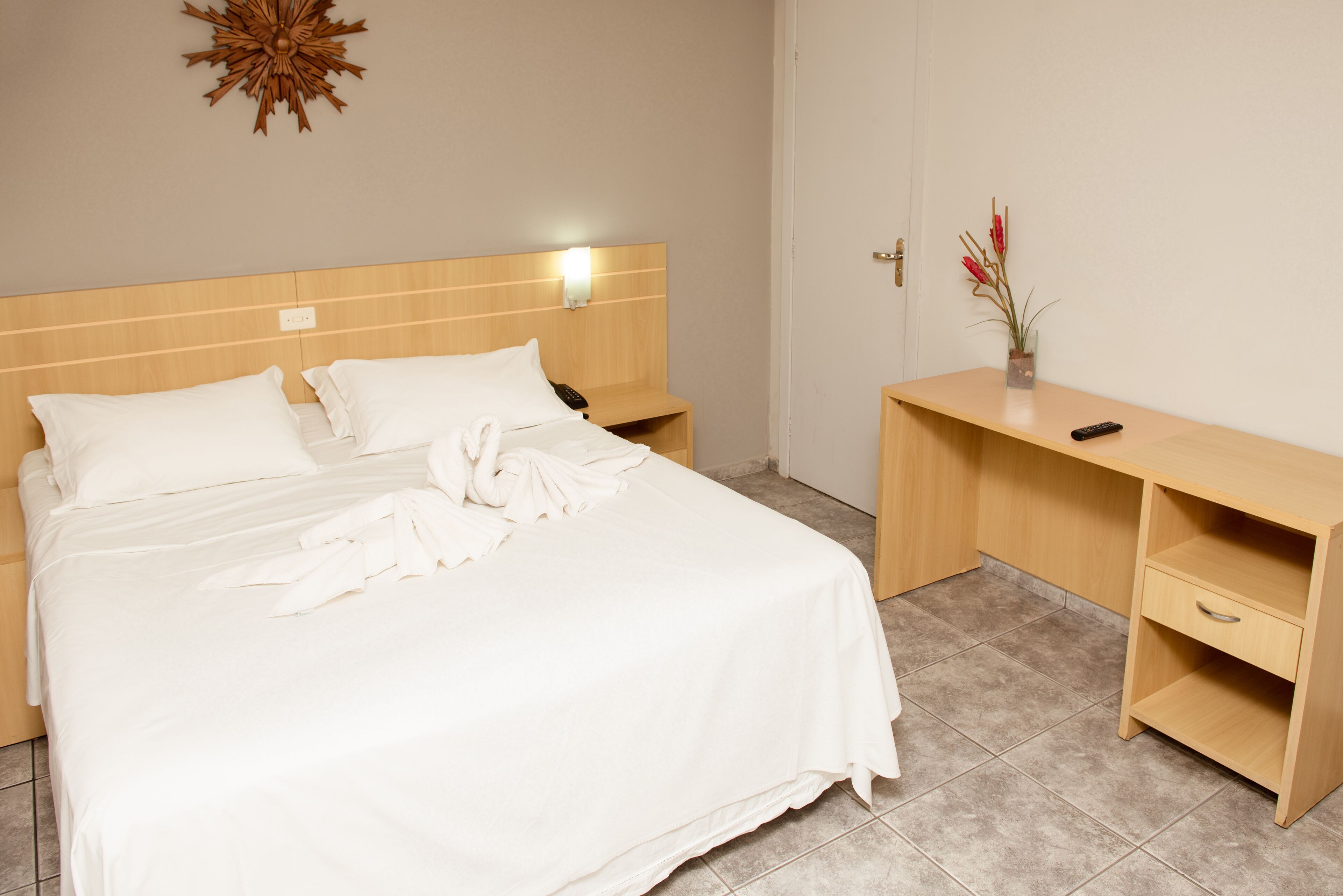 Una cama bien hecha con ropa blanca, un cabecero de madera, una mesita de noche con una lámpara y un pequeño escritorio con un jarrón de flores.