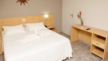 Una cama bien hecha con ropa blanca, un cabecero de madera, una mesita de noche con una lámpara y un pequeño escritorio con un jarrón de flores.