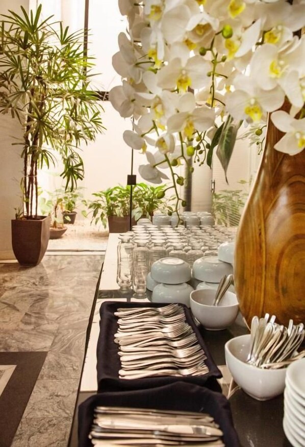 A buffet setup with neatly arranged plates, bowls, and utensils on a marble-tiled floor, surrounded by potted plants and a large vase. - River Palace Hotel (Divinopolis)