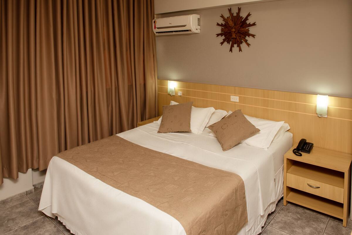 A hotel room with a bed, a nightstand, an air conditioner, and a decorative wall piece.
