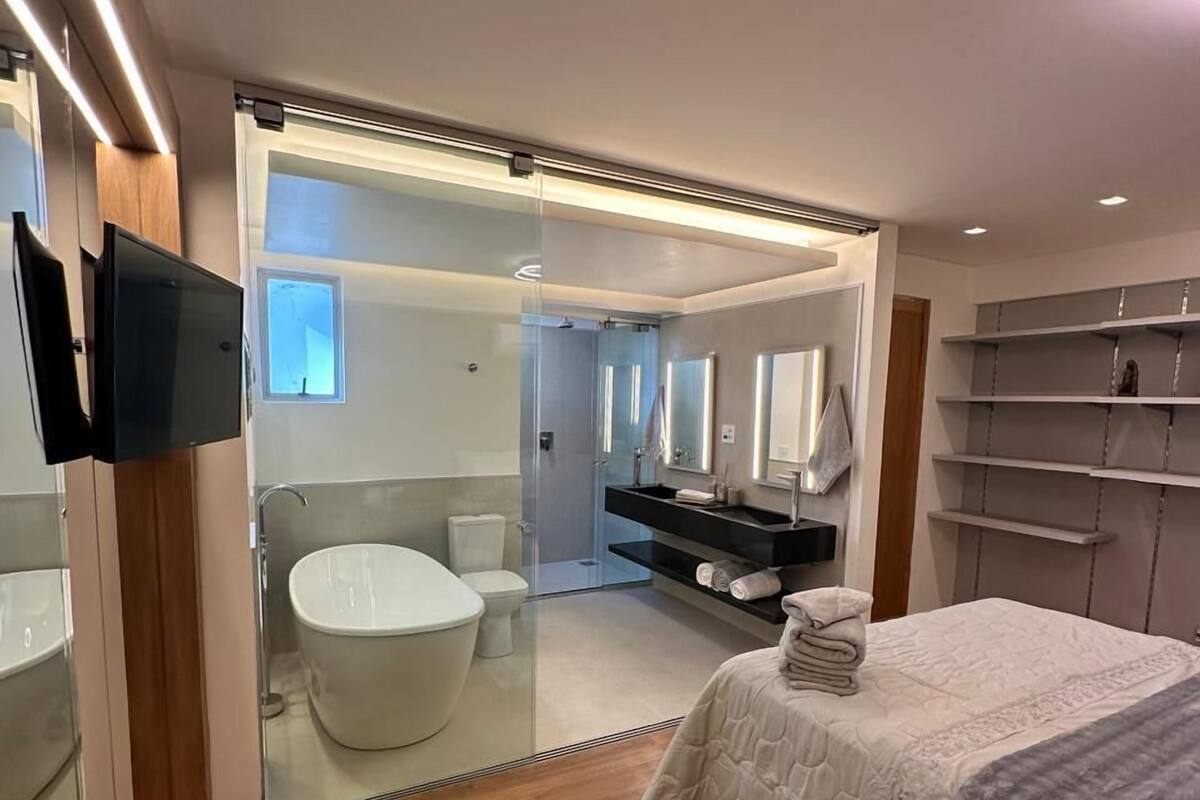 A modern bedroom with a large bed, a glass-enclosed bathroom, and a TV mounted on the wall.